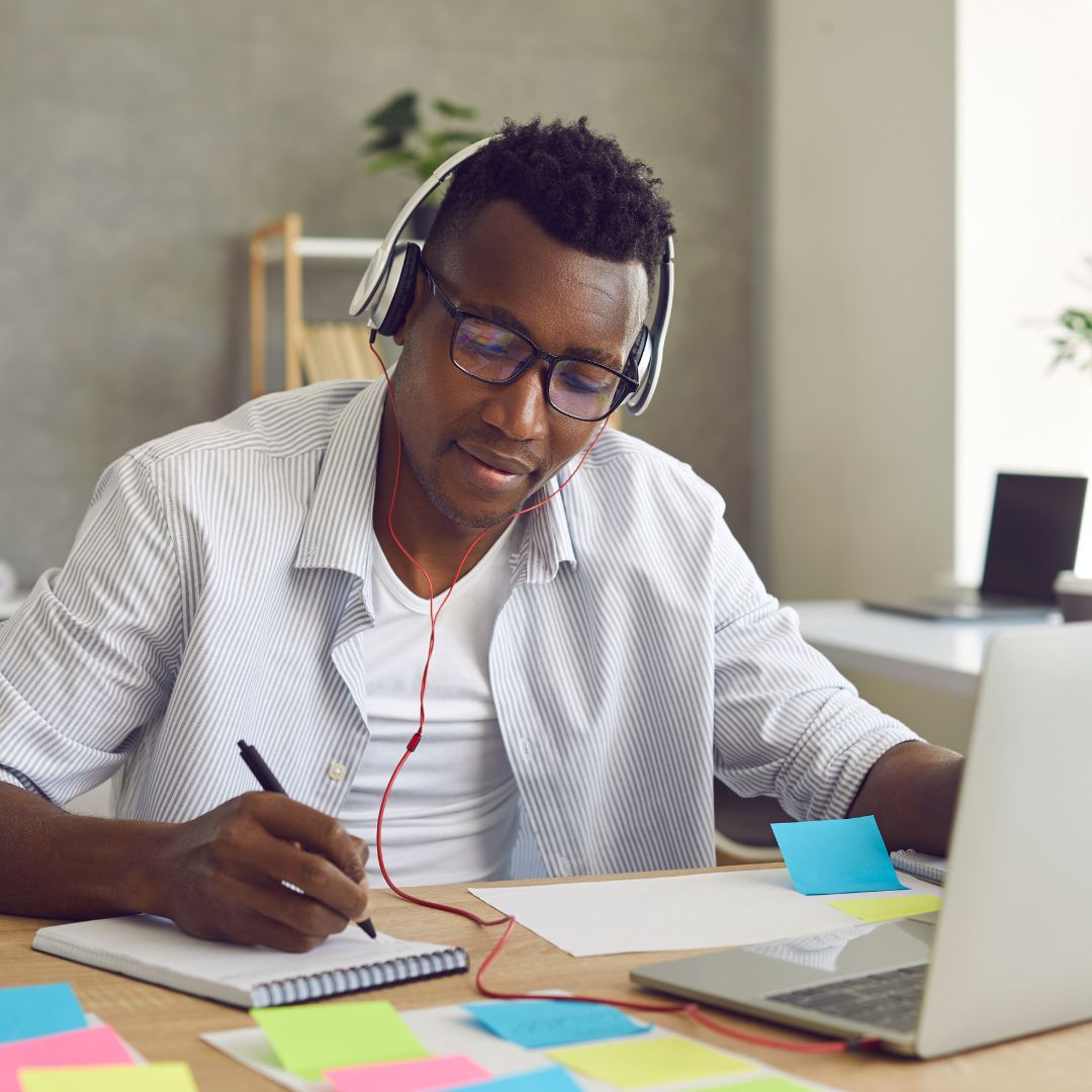 man at desk taking notes while using laptop with headphones on